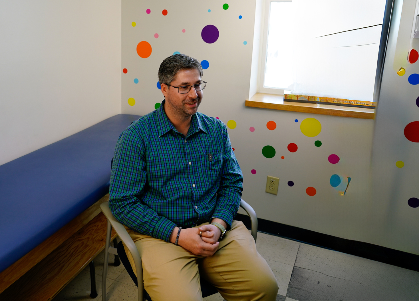 A photograph of Dr. Adam Gicewicz sitting in a pediatric exam room with lots of colored dots on the wall behind him.