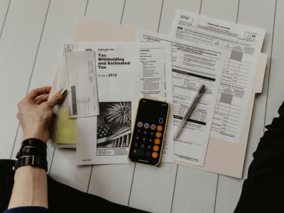 A photograph of a hand holding some of a series of papers which appear to be tax related, a calculator and a pen rest on the papers