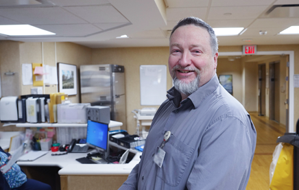 A photograph of a male nurse standing near an office counter, looking at the camera