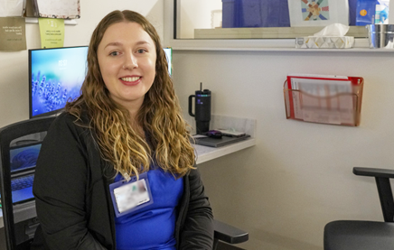 A photograph of a young woman Nurse, sitting in front of a desk turning to look at the camera and smiling.