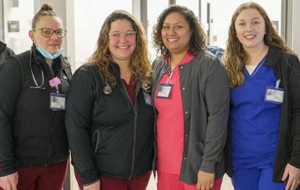 A photograph of four nurses standing side by side looking at the camera, all smiling big smiles.