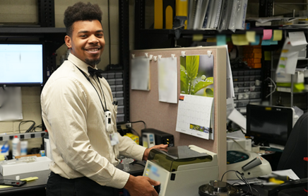 A photograph of a biomedical tech standing in a crowded office space, turning away from a piece of medical equipment to look at the camera