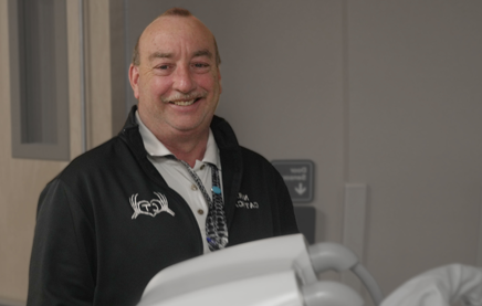 A photograph of a radiology tech standing on the fas side of some radiological equipment, smiling at the camera.