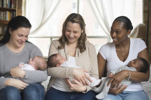 A photograph of a group of three mothers, both black and white, sitting on a couch, with their babies.