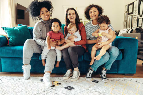 A photograph of a group of three mothers, both black and white, sitting on a blue couch with their babies.