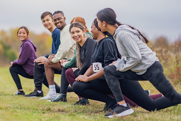 A large group of adolescents are seen standing side-by-side outside as they stretch out their legs and warm up before a cross country race. They are each wearing athletic wear and numbered race bibs.
