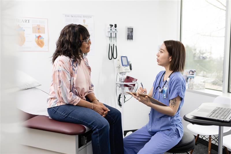 Patient sitting on an exam table engaged in conversation with a healthcare provider sitting nearby.